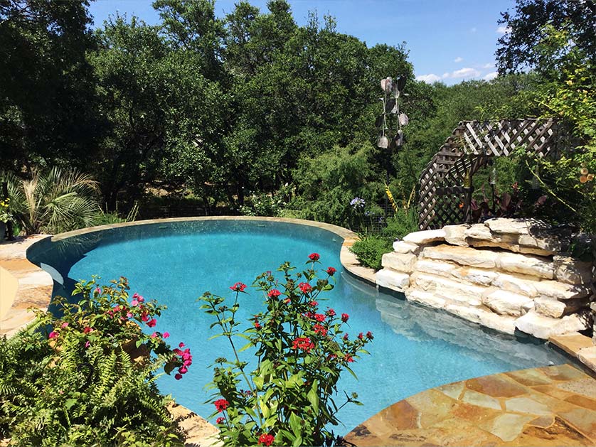 Custom pool built into natural backdrop of trees and landscaping. Natural stone waterfall and floral landscaping surround the custom shaped pool.