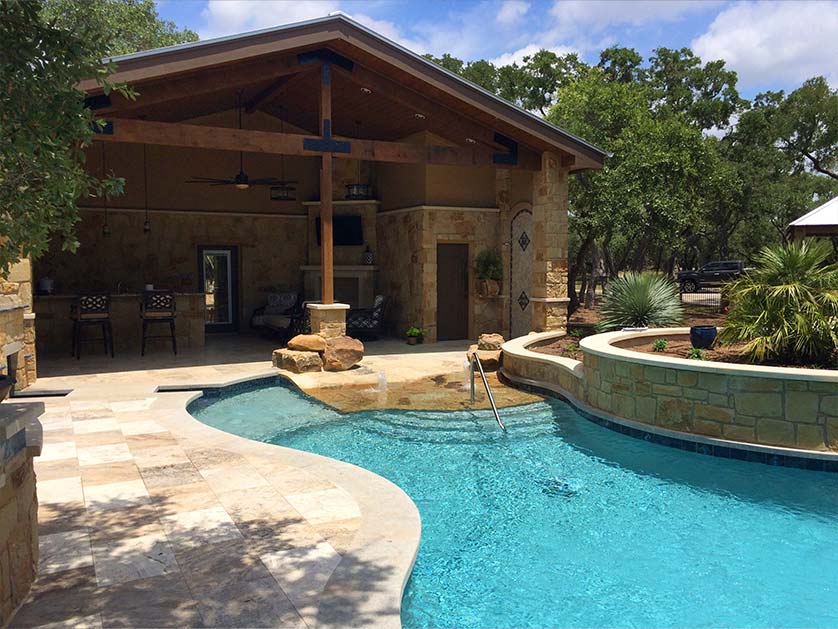 Custom outdoor patio area covered by extended roofline from the house. Zero entry pool with sitting area in the shallow water and adjacent landscaped planting area.
